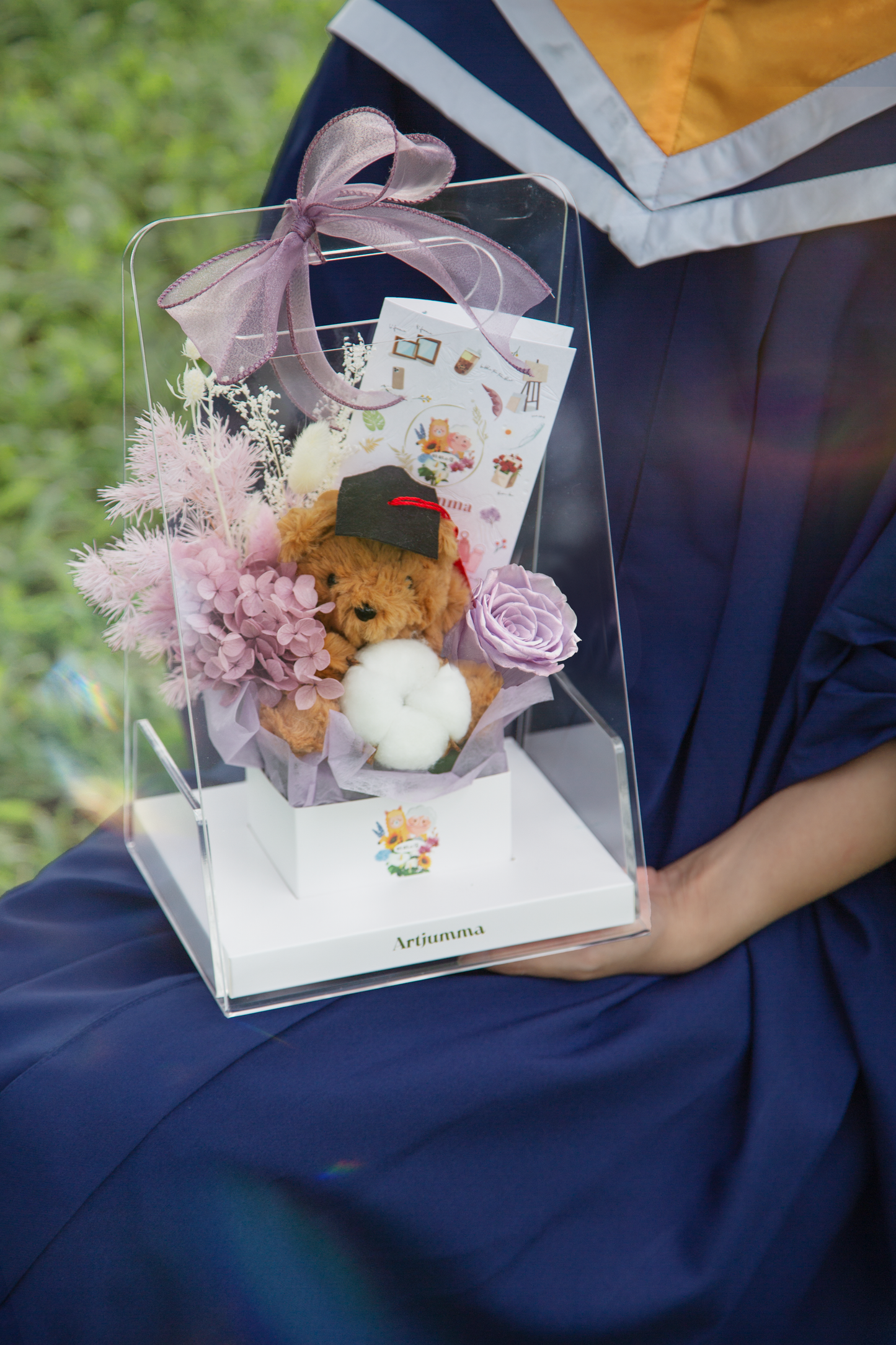 Girl's hands holding graduation teddy bear and preserved purple flowers gift box with purple ribbon, with gift card inside.